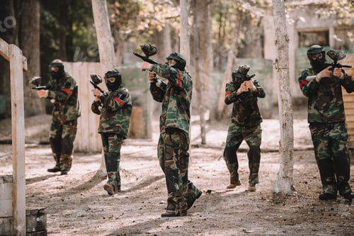 Preview: paintball team in uniform and protective masks shooting by paintball guns outdoors