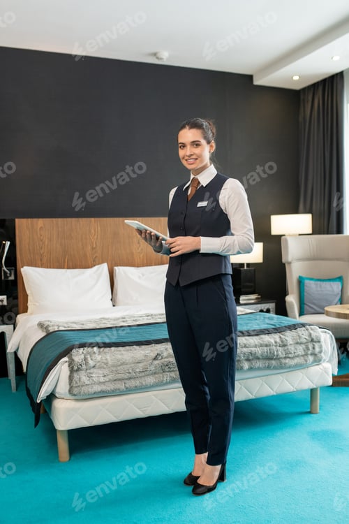 Preview: Happy young elegant female worker of hotel standing in front of camera against bed and using tablet