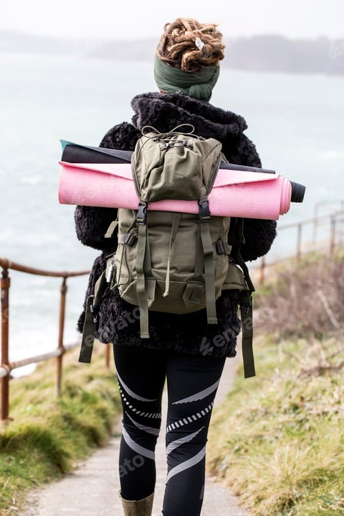 Preview: Rear view of young woman with brown hair and dreadlocks carrying backpack, walking along coastal