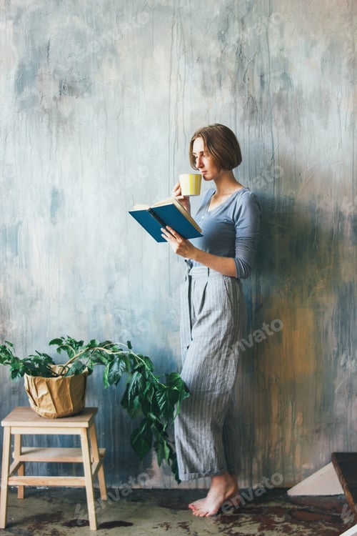 Preview: Young woman with yellow cup standing against wall and reading book in the art work studio
