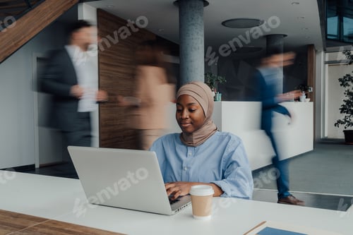Preview: Focused black muslim woman working on laptop in busy office
