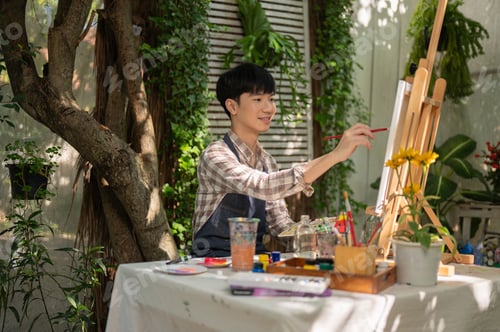 Preview: Young handsome Asian man is focusing on painting on a canvas easel in his beautiful green garden.