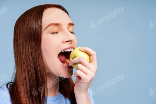 Preview: Closeup of gorgeous red haired female wearing braces, enjoying apple, isolated on a blue background