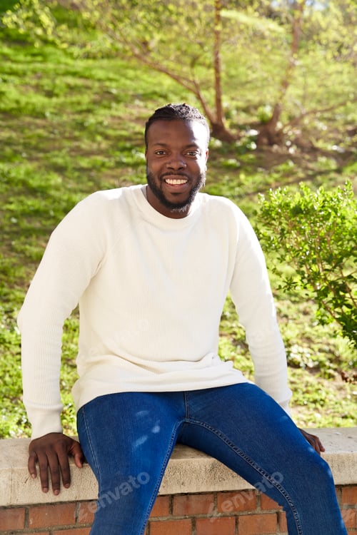 Preview: Young African American man smiling sitting on a wall. Happy Latino man.