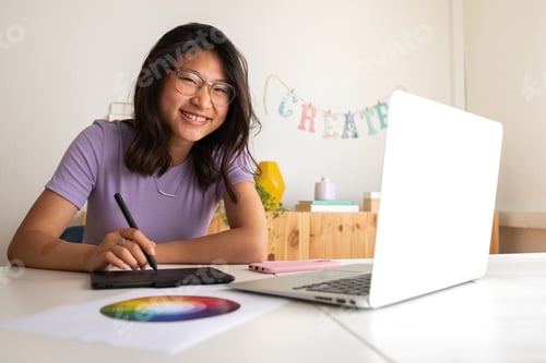 Preview: Asian female graphic design student working at home using tablet and laptop looking at camera.
