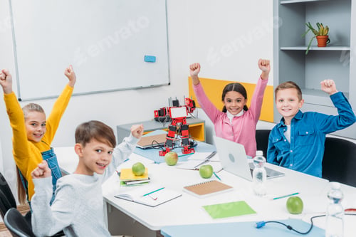 Preview: schoolchildren joying and raising hands at desk in stem education class