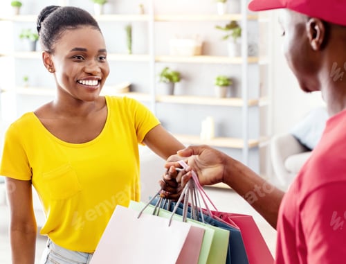 Preview: Happy Black Woman Receiving Shopper Bags From Courier Standing Indoor