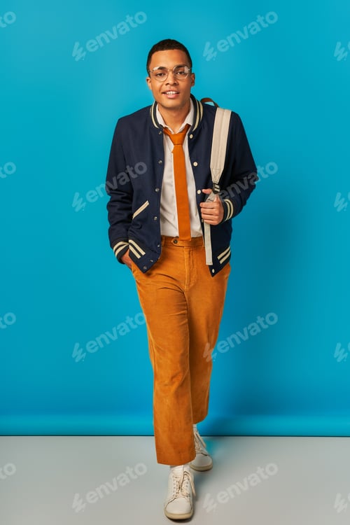 Preview: stylish african american student in jacket and orange pants walking with hand in pocket on blue