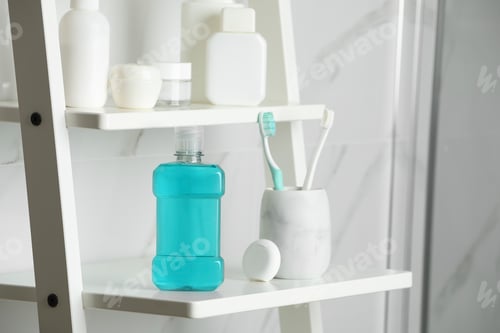 Preview: Bottle of mouthwash, toothbrushes and dental floss on white shelf in bathroom
