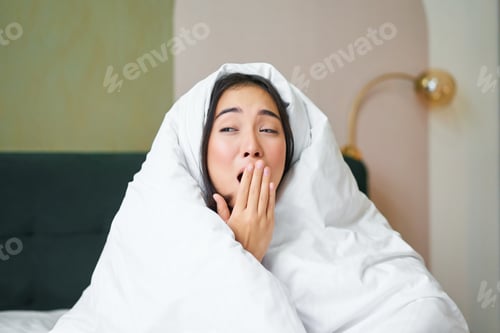 Preview: Close up of funny sleepy girl, asian woman yawns after waking up early morning, covers herself with