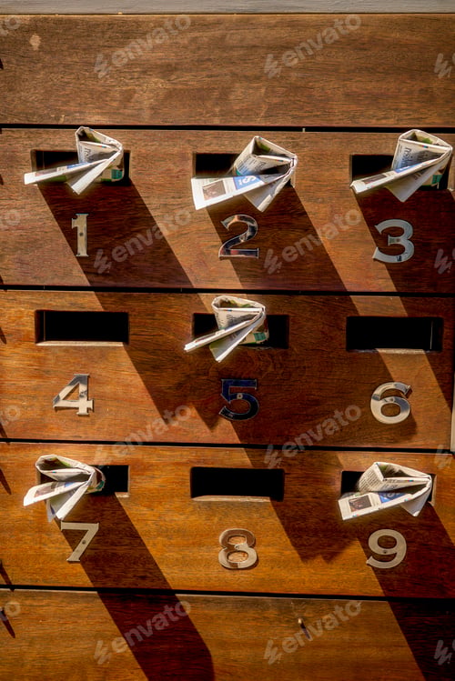 Preview: A vertical array of wooden mailboxes with numbers from 1 to 9, each containing rolled-up newspapers.