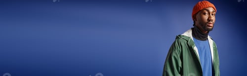 Preview: stylish african american man in knitted hat and trendy winter attire posing on blue backdrop, banner