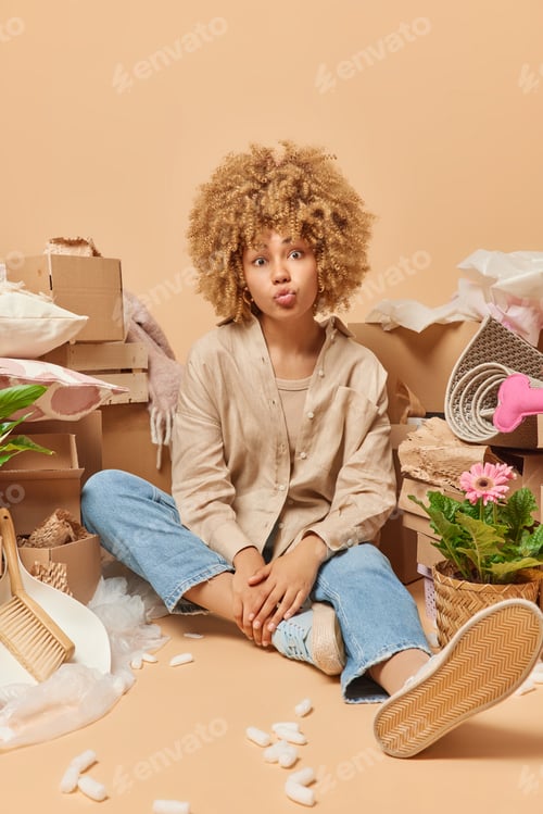 Preview: Vertical shot of curly haired young woman keeps lips folded sits on floor near carton boxes dressed