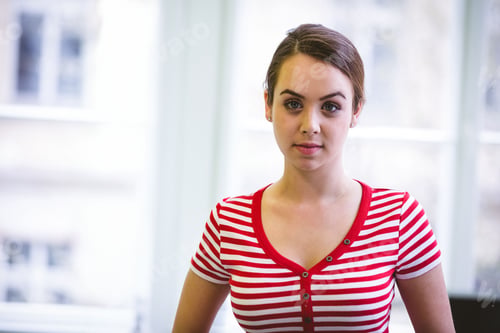 Preview: Woman standing facing camera by floor-to-ceiling windows in bright studio wearing striped shirt