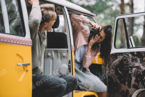 Preview: girlfriend taking photo of her boyfriend on camera while sitting in retro styled minivan