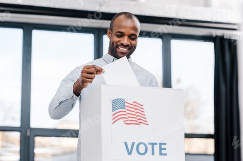Preview: happy african american man voting and putting ballot in box with flag of america