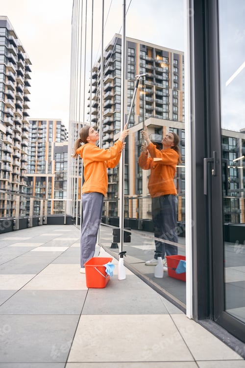 Preview: Young employee cleaning company in warm hoodie washes mirrored windows