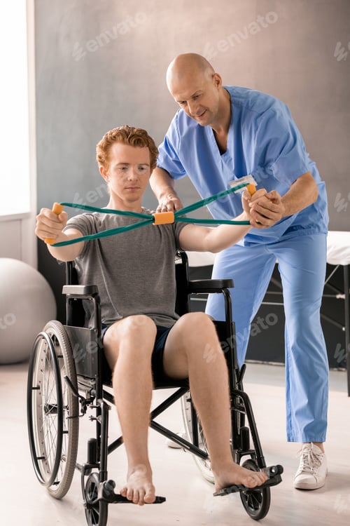 Preview: Mature clinician helping young patient in wheelchair while supporting his hand