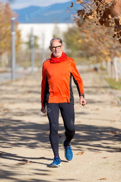 Preview: Front view of a senior man in sport clothes jogging in a city park in a sunny day