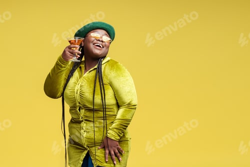 Preview: Happy stylish African American woman holding glass with cocktail isolated on yellow. Birthday party
