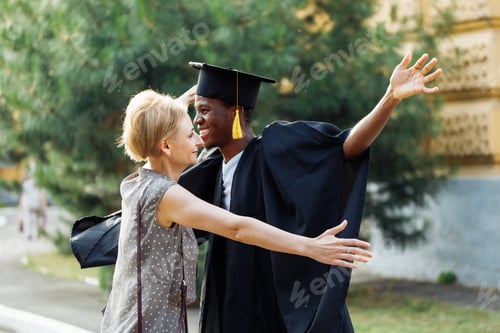 Preview: Portrait of smiling afro american student in black graduation mantle and hat open arms wide to hug