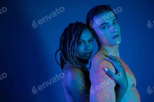 Preview: sexy interracial couple in colorful neon body paint looking at camera on blue background with cyan