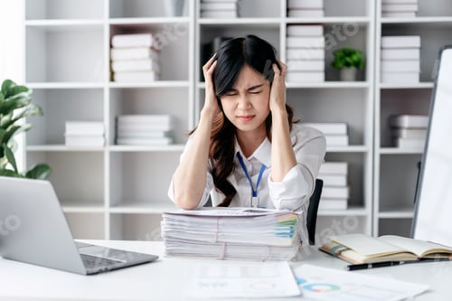 Preview: A woman is sitting at a desk with a laptop and piles of papers