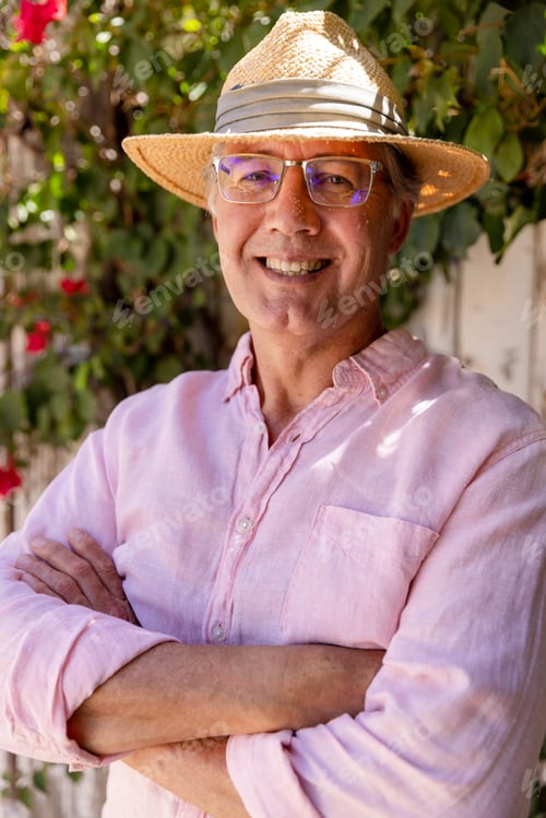 Preview: Smiling caucasian senior man wearing hat and eyeglasses with arms crossed standing against plants