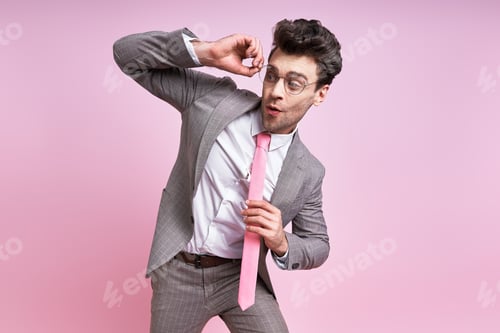 Preview: Surprised man in full suit adjusting his eyeglasses while standing against pink background