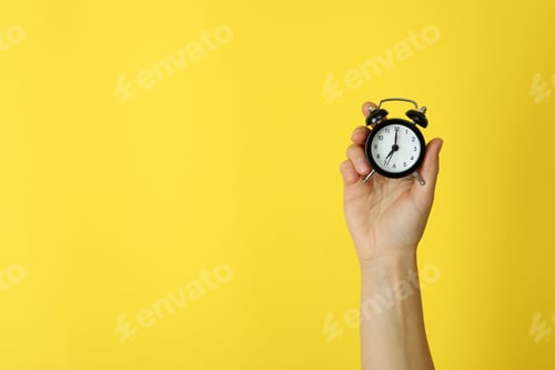 Preview: Female hand holds alarm clock on yellow background