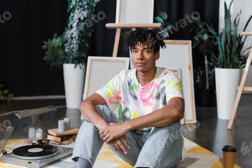 Preview: African american artist looking at camera near vinyl player and books in studio