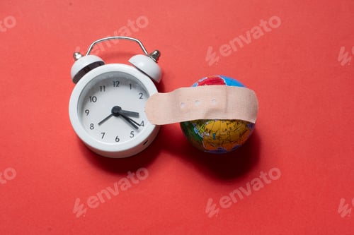 Preview: High angle shot of an alarm clock and small globe with a bandaid on a red surface