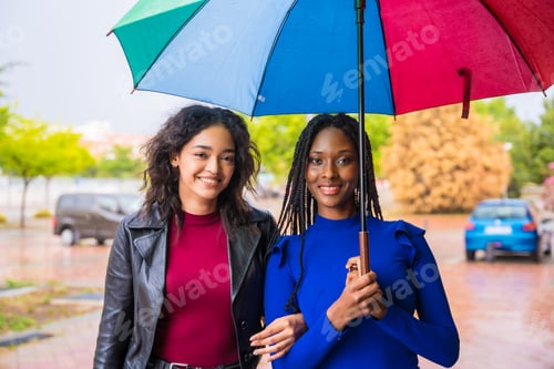 Preview: Portrait of laughing multi-ethnic female friends with an umbrella in the rain in a city in spring