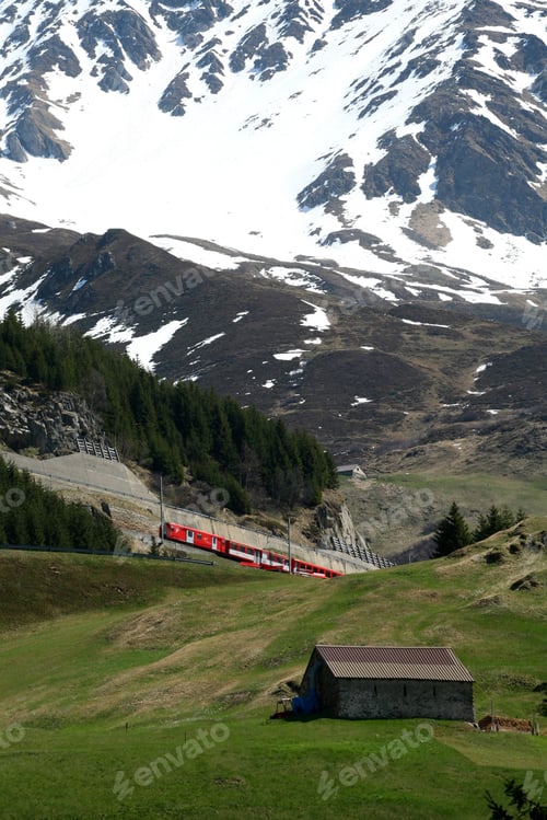 Preview: Vibrant red train travels through a lush green valley with snow-capped mountains in the background.