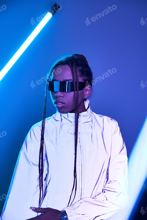 Preview: african american girl with dreadlocks posing in trendy jacket and sunglasses on blue with neon light