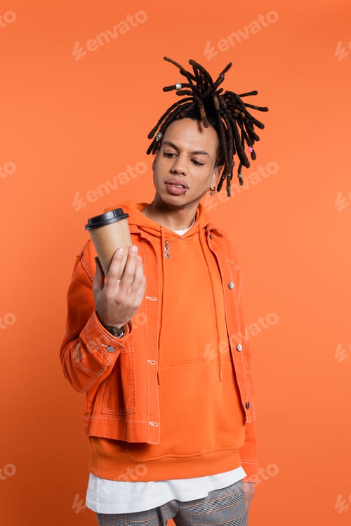 Preview: tattooed and multiracial man with dreadlocks looking at paper cup with coffee to go isolated on