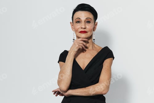 Preview: Elegant senior woman in black dress and earrings striking a pose with hand on chin before camera
