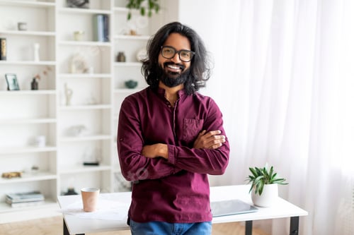 Preview: Smiling Indian Man Standing With Folded Arms Near Desk At Home Office