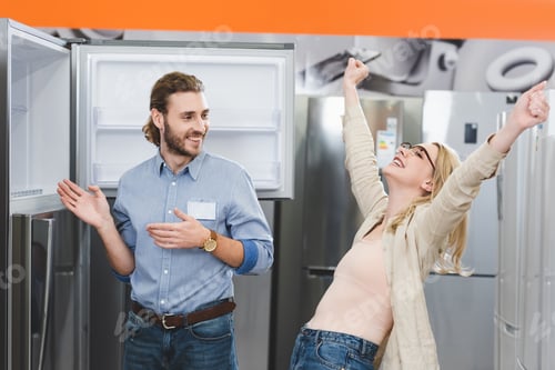 Preview: consultant pointing with hands at fridge and smiling woman showing yes gesture in home appliance