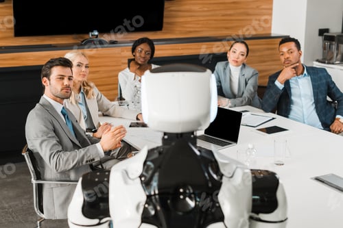 selective focus of young multicultural businesspeople looking at robot while sitting in conference