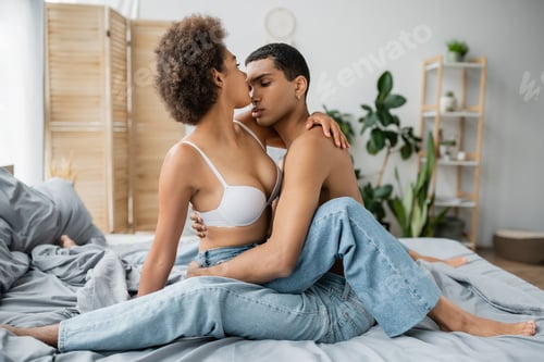 Preview: young african american woman with sexy body near shirtless boyfriend sitting on bed in jeans