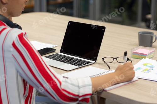 Preview: Midsection of biracial young businesswoman writing in diary by blank screen laptop with copy space