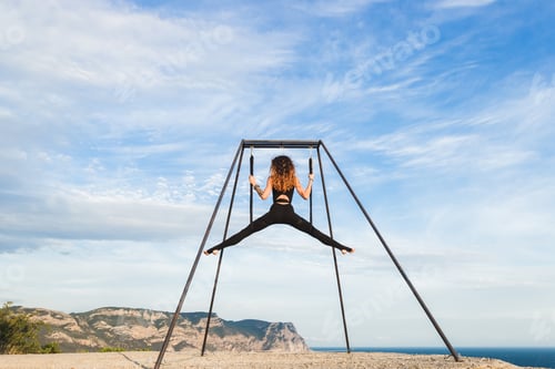 Preview: Woman practicing fly-dance yoga poses in a hammock on the coast with views of the mountains