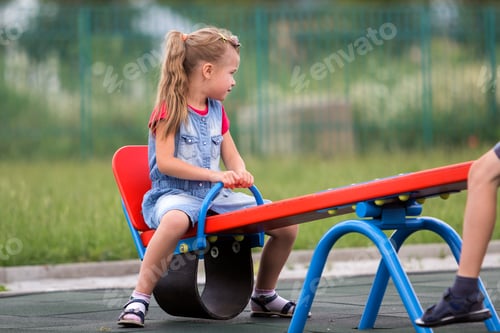 Preview: Small cute young blond child girl sits on see- saw swing on warm sunny day
