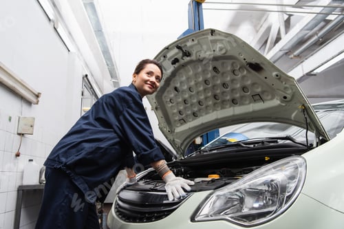 Preview: Smiling mechanic in uniform standing near car with open hood