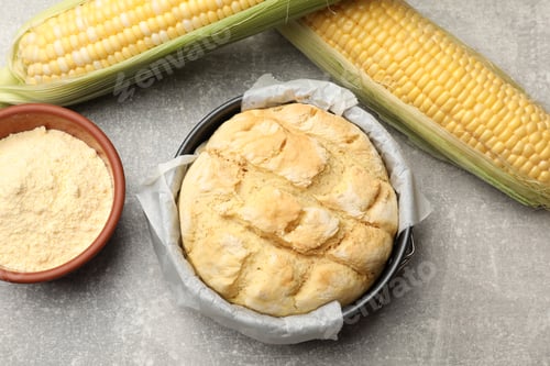 Preview: Freshly baked cornbread in cake pan, cobs and flour on grey textured table, flat lay
