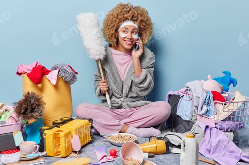 Preview: Indoor shot of housewife has rest after cleaning house makes phone call holds dusting brush
