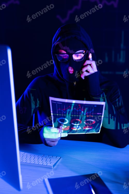 Preview: Hacker in balaclava talking on smartphone and holding paper near computer on black background