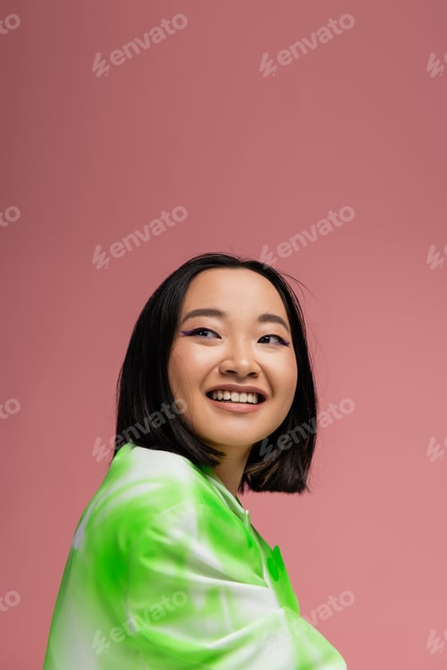 Preview: portrait of brunette asian woman in stylish clothes and makeup looking away isolated on pink