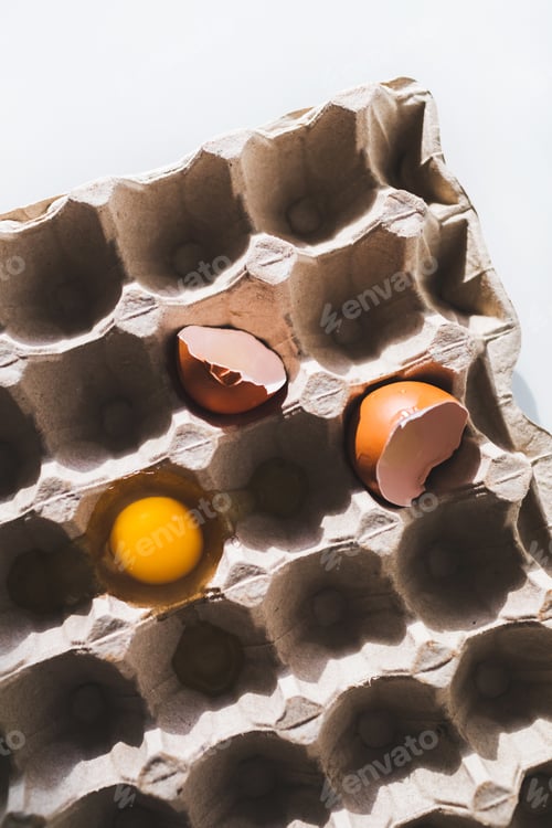 Preview: Yellow yolk and cracked eggshell in carton egg box. Easter vertical background and concept.